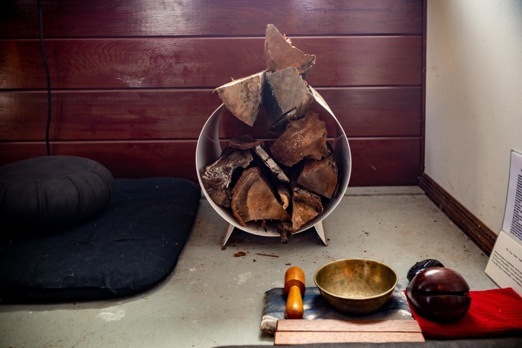 A round piece of metal holds chopped firewood. In front of it is the moktak, and small kesu bell which are both used for time keeping during a sit.
